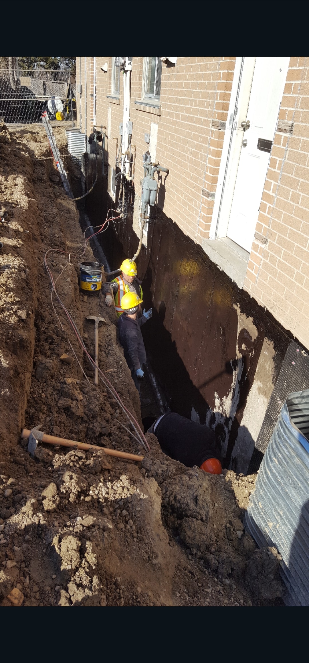 Foundation Excavation — Three Workers in Full PPE Applying Waterproofing